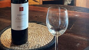 A bottle of wine on a woven mat with a glass beside it, set on a wooden table in a kitchen with a brick backsplash.