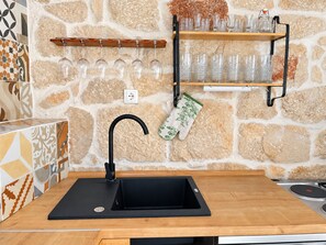 Une cuisine avec un évier noir, un plan de travail en bois, un mur en pierre et une étagère où sont rangés des verres.