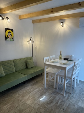 Une pièce avec un canapé vert, une table à manger blanche avec des chaises, un tableau au mur et un luminaire au plafond.
