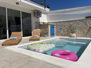 A swimming pool with lounge chairs and inflatable rings, a stone wall, and a blue door.