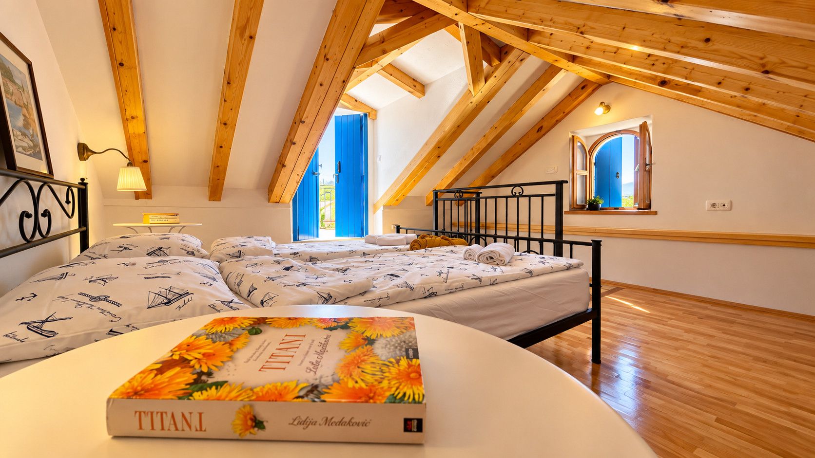 A bedroom with a sloped ceiling, wooden beams, a bed with patterned bedding, a bedside table, and a window with blue shutters.