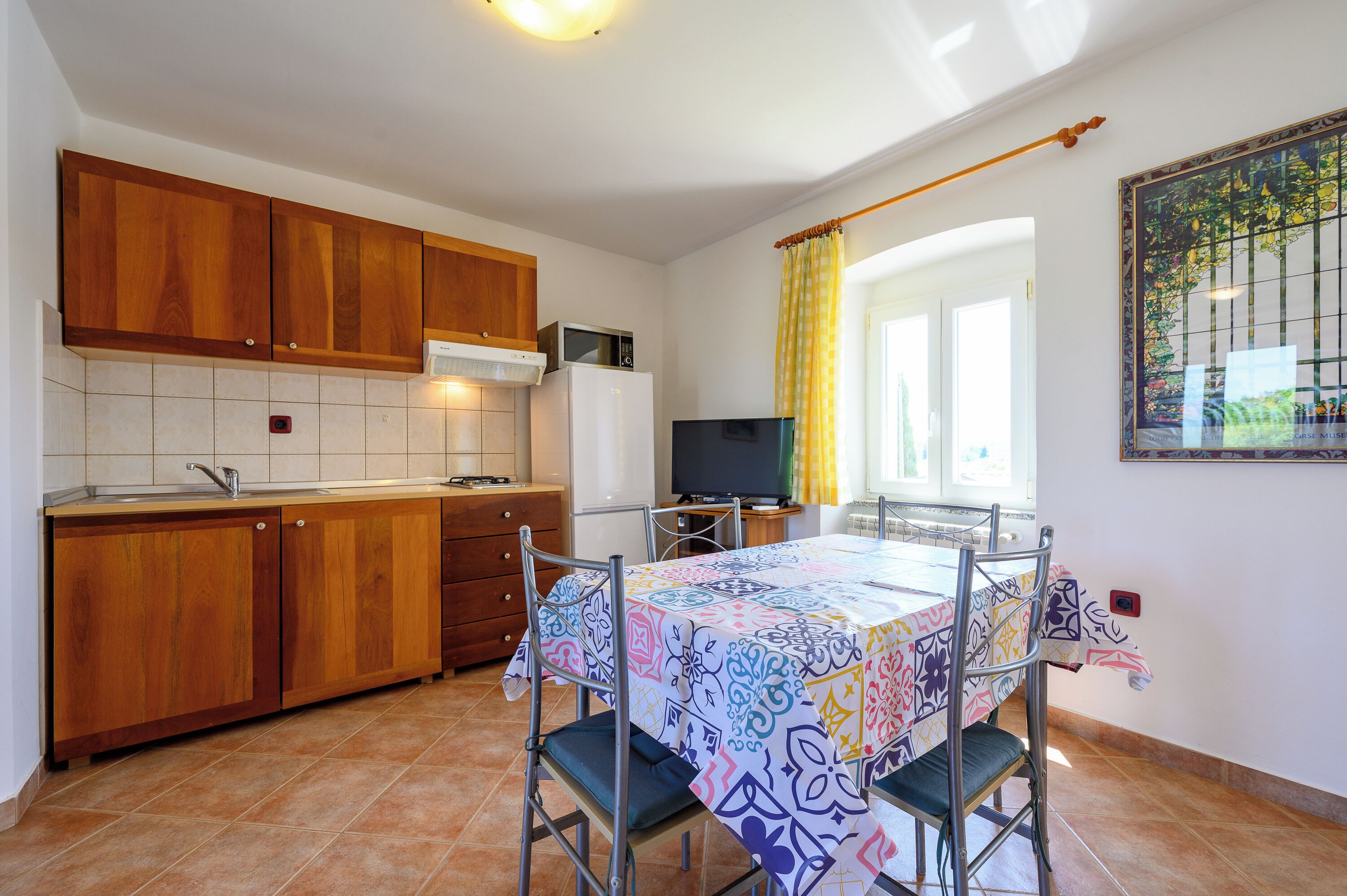 Eine kleine Küche mit Holzschränken, einer Mikrowelle und einem Kühlschrank. Ein Esstisch mit bunten Tischdecken und Stühlen lädt zum Verweilen ein. Ein Fernseher steht auf einem Beistelltisch neben dem Fenster, das mit gelben Vorhängen drapiert ist.