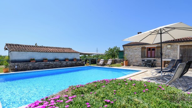 Une piscine bordée d’un muret en pierre, un espace détente avec des fauteuils et une pergola dotée d’un parasol.