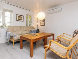 A living room with a sofa, a wooden table, and two wicker chairs. There is a window with curtains, an air conditioning unit on the wall, and framed pictures on the wall. - 6 Person Holiday Home in Horslunde (Horslunde)