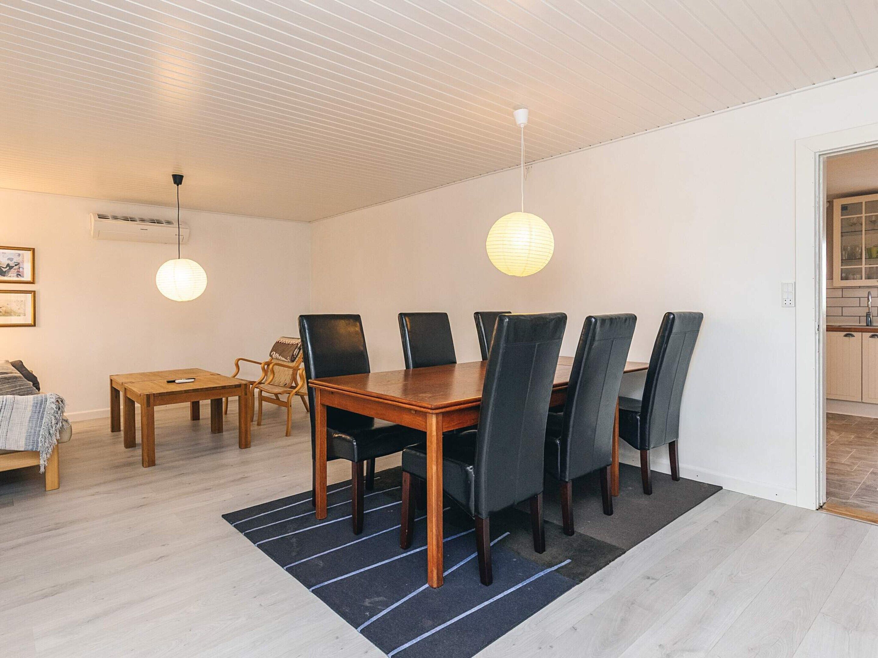 A dining area with a wooden table and chairs, a black rug, and pendant lights.