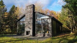 Una cabaña de madera moderna con ventanas amplias y una chimenea, rodeada de un bosque.