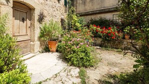 A courtyard with a stone building, a wooden door, potted plants, and flowering bushes. - LA FONTAINE – Peaceful retreat in medieval Tourrettes-sur-Loup (Tourrettes-sur-Loup)
