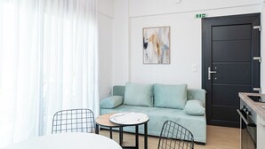 Uma sala de estar moderna com um sofá azul claro, uma mesa redonda pequena e uma cadeira. Há um quadro na parede e uma porta preta com puxador.