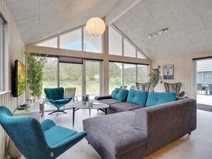 Ein modernes Wohnzimmer mit einem Ecksofa, türkisfarbenen Sesseln, einem Couchtisch und großen Fenstern mit Blick ins Freie.