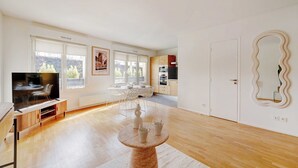 Un salon moderne avec un téléviseur, une table à manger et des chaises. On aperçoit une cuisine à l’arrière-plan, avec des placards et un réfrigérateur.