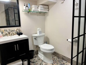 Un baño con inodoro blanco, lavamanos con artículos de aseo, un espejo y un estante con toallas.