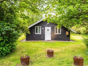 A small, dark wooden cabin with a white door and two windows, surrounded by lush greenery. - 4 Person Holiday Home in Hemmet (Hemmet)