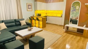 A living room with a sofa, coffee table, and ottomans. There is a kitchen area with a yellow backsplash and white cabinets. A painting is on the wall, and a small white shelf is on the right side. - city center 1-bedroom apartment with WiFi and AC in delightful Shkodër (Shkodër)