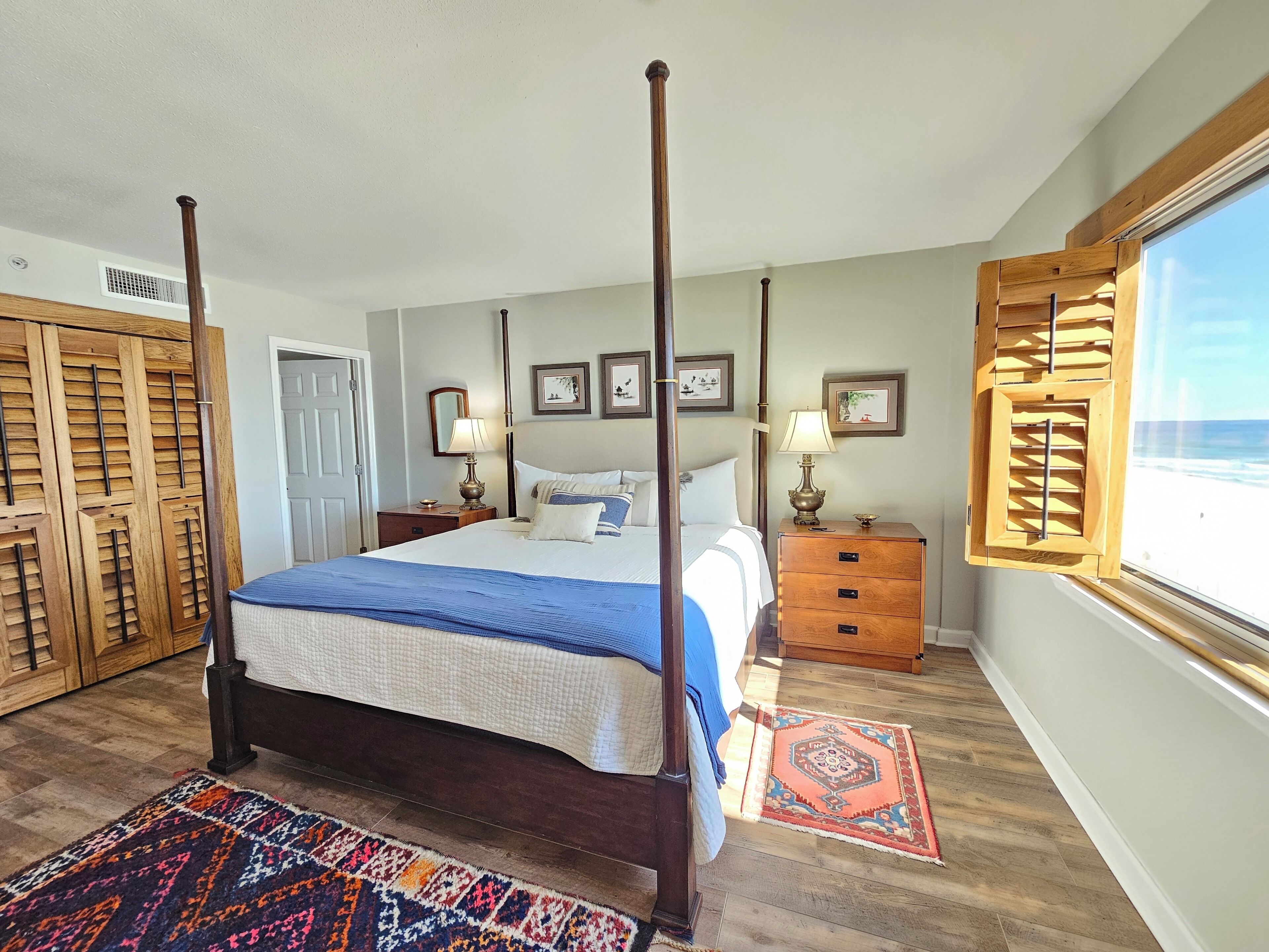 A four-poster bed with a blue and white bedspread, wooden nightstands with lamps, a wooden wardrobe, and a large window with a beach view.