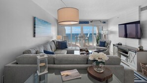 A modern living room with a grey sofa, a coffee table with a book and flowers, a TV, and a balcony with a view of the sea. - Sandy Key 816: 2 BR/2 BA Perdido Key Beachfront Condo, On-Site Rental Office, Sleeps 6 (Pensacola)