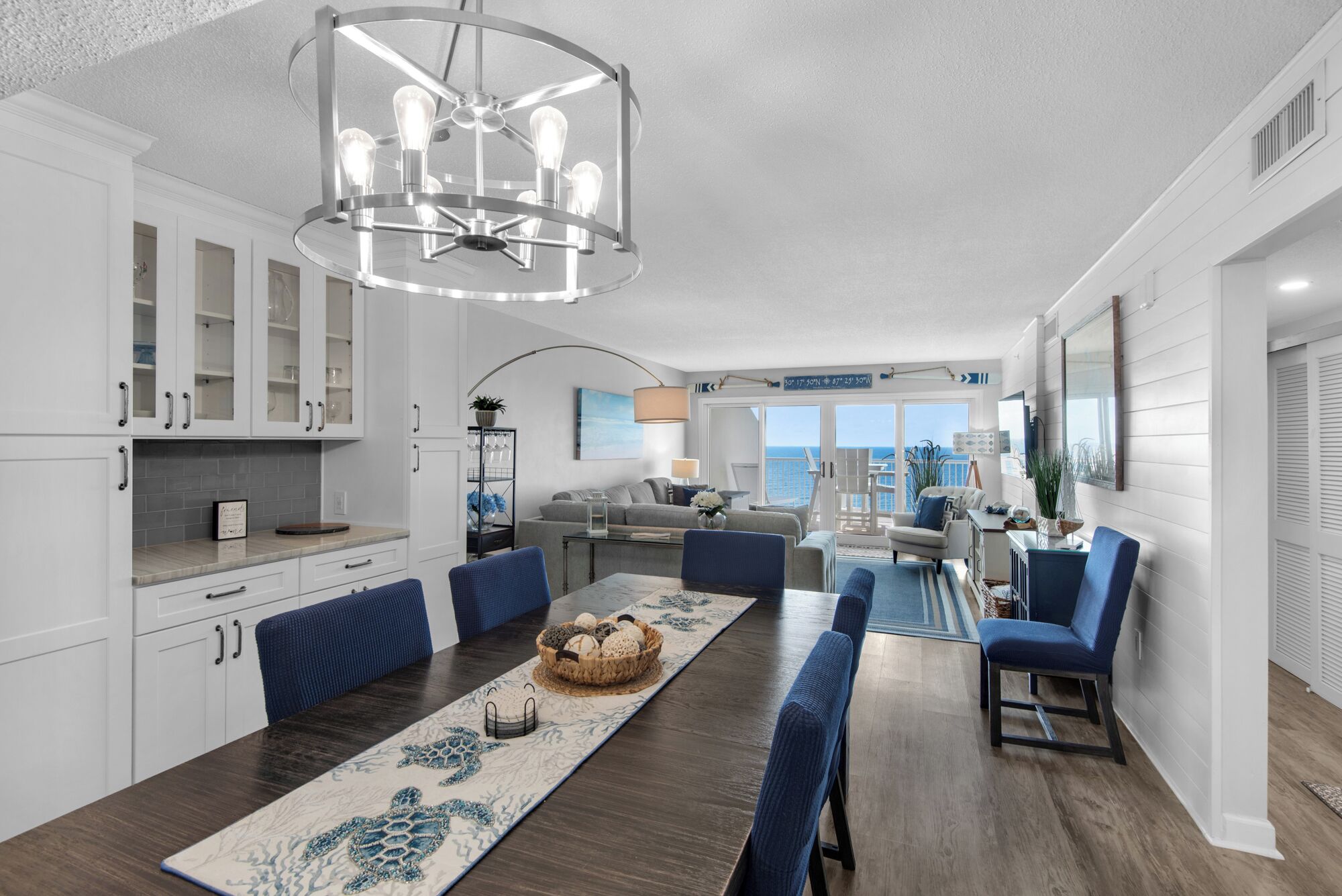 A modern kitchen with white cabinetry, a dining area with a long table, and a view of the ocean through large windows.