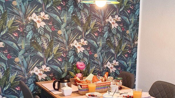 Een eethoek met een houten tafel, stoelen en behang met een tropisch motief.