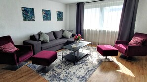 Een moderne woonkamer met een grijze bank, twee fauteuils, een salontafel en een vloerkleed met een patroon. Boven de bank hangen drie kunstwerken.