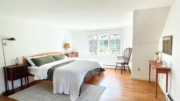 Ein Schlafzimmer mit Bett, Nachttisch, Lampe, Stuhl und einem Fenster mit Blick auf Bäume.