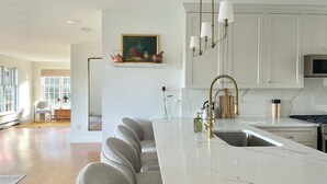 Una cocina moderna con encimera de mármol, grifo dorado y armarios blancos. Se aprecia un cuadro en la pared y una silla al fondo.