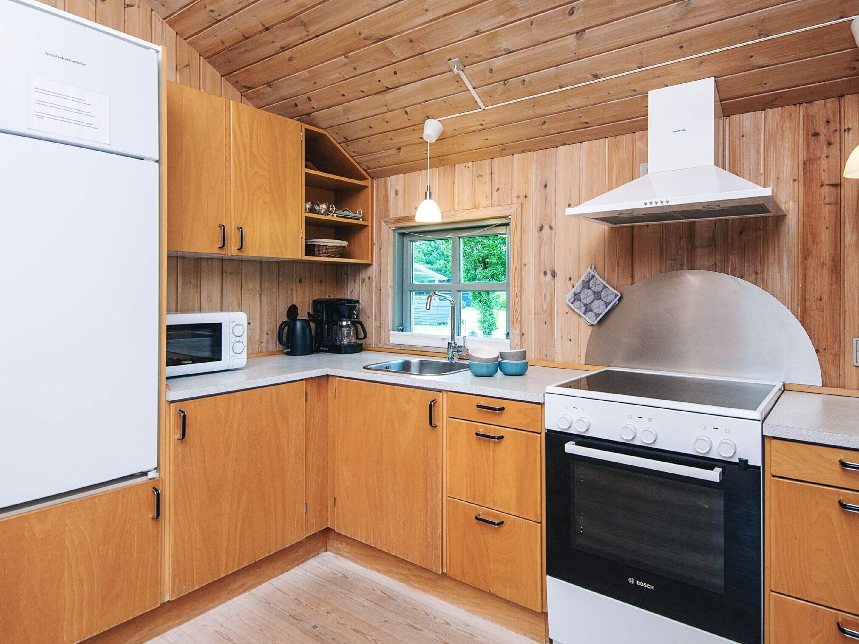 A compact kitchen with wooden cabinets, a Bosch oven, a microwave, a coffee maker, and a window providing natural light.