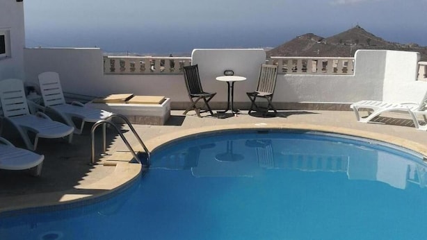 Une piscine entourée de chaises longues et d’une table, le tout bordé d’un mur blanc d’où l’on peut admirer une montagne.