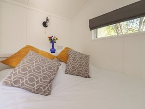 Un dormitorio con una cama, almohadas, un florero con flores, una lámpara de pared y una ventana.