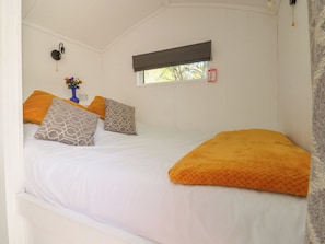 A bed with white bedding and multiple pillows, a window with a blind, a wall-mounted light fixture, and a small vase with flowers. - Butterfly Hut 2 (Preston)