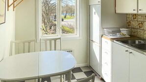 A kitchen with a white table and chairs, a window with a view of trees and a road, and white cabinets with a tiled backsplash. - 4 Star Holiday Home in Ronneby (Ronneby)