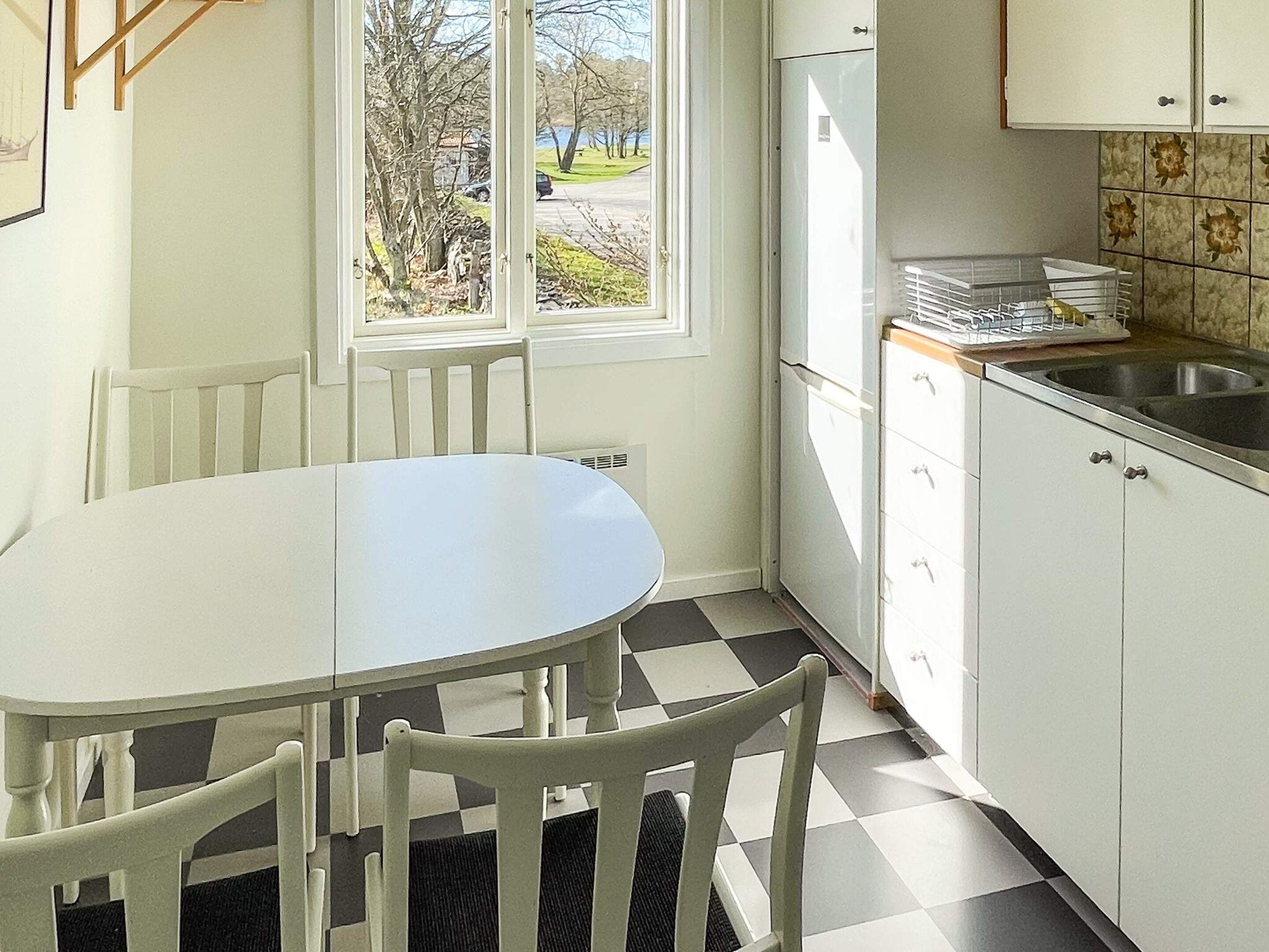 Una cocina con suelo a cuadros, armarios blancos, una ventana con vistas a árboles y una carretera, y una mesa con sillas.