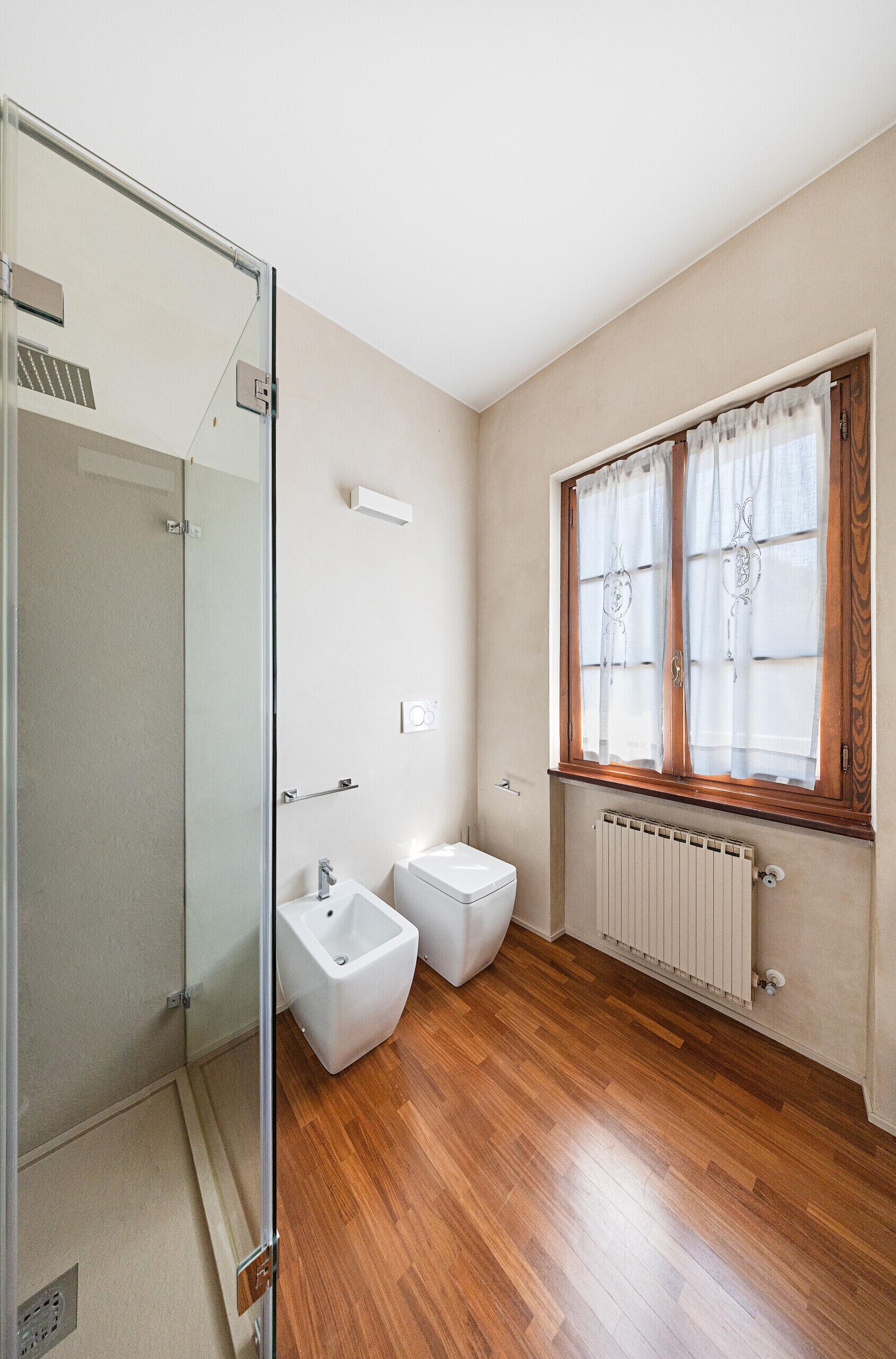 Een badkamer met een douchecabine van glas, een bidet, een toilet en een raam met gordijnen.
