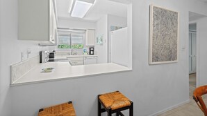 A modern kitchen with white cabinetry, a large mirror, and a countertop. There are two stools with woven seats. - Pelican Harbour, Beach Club T2 Condo by RVA (Longboat Key)