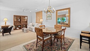 A dining area with a round table, chairs, a chandelier, a ceiling fan, and a large mirror on the wall. - Pelican Harbour, Beach Club T2 Condo by RVA (Longboat Key)