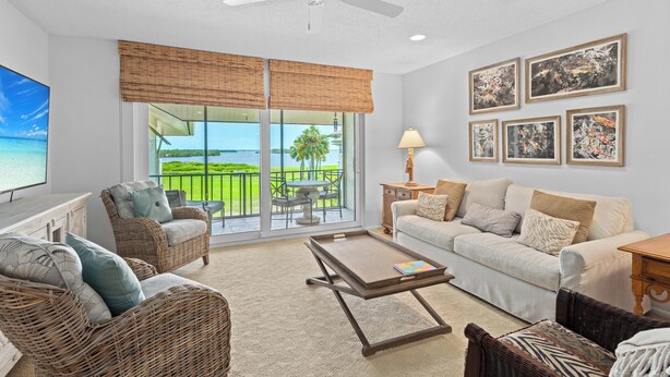A spacious living room with a large window, wicker furniture, a ceiling fan, and artwork on the walls. - Pelican Harbour, Beach Club T2 Condo by RVA (Longboat Key)
