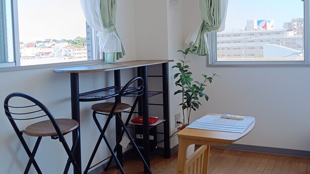 A room with a wooden floor, a small table with a chair, a potted plant, and a window with green curtains.