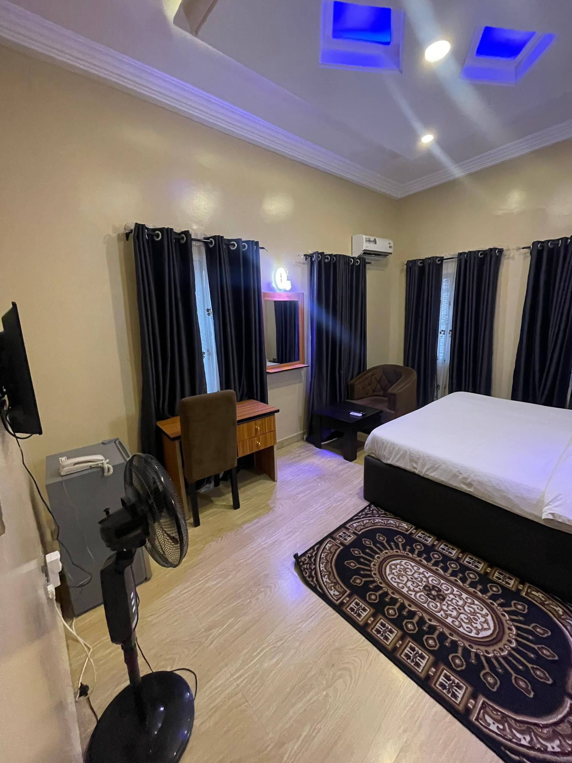 A hotel room with a bed, a desk, a chair, a fan, and a large rug.