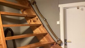 Un escalier en bois avec une rampe en corde, menant à une porte dotée d’un interrupteur et ornée d’un petit objet décoratif sur le mur.