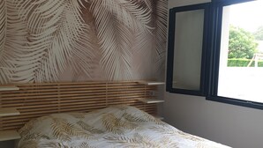 A bedroom with a bed, a wooden headboard, a wall-mounted mirror, and a window with a view of greenery outside.