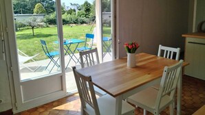 A dining area with a wooden table and white chairs, a vase with red flowers, and a view of a garden through glass doors.