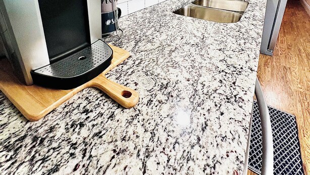 A kitchen with white cabinets, a granite countertop, a coffee maker, a sink, and a wooden cutting board.