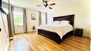 A bedroom with a wooden floor, a bed with white bedding, a ceiling fan, and a window with curtains.