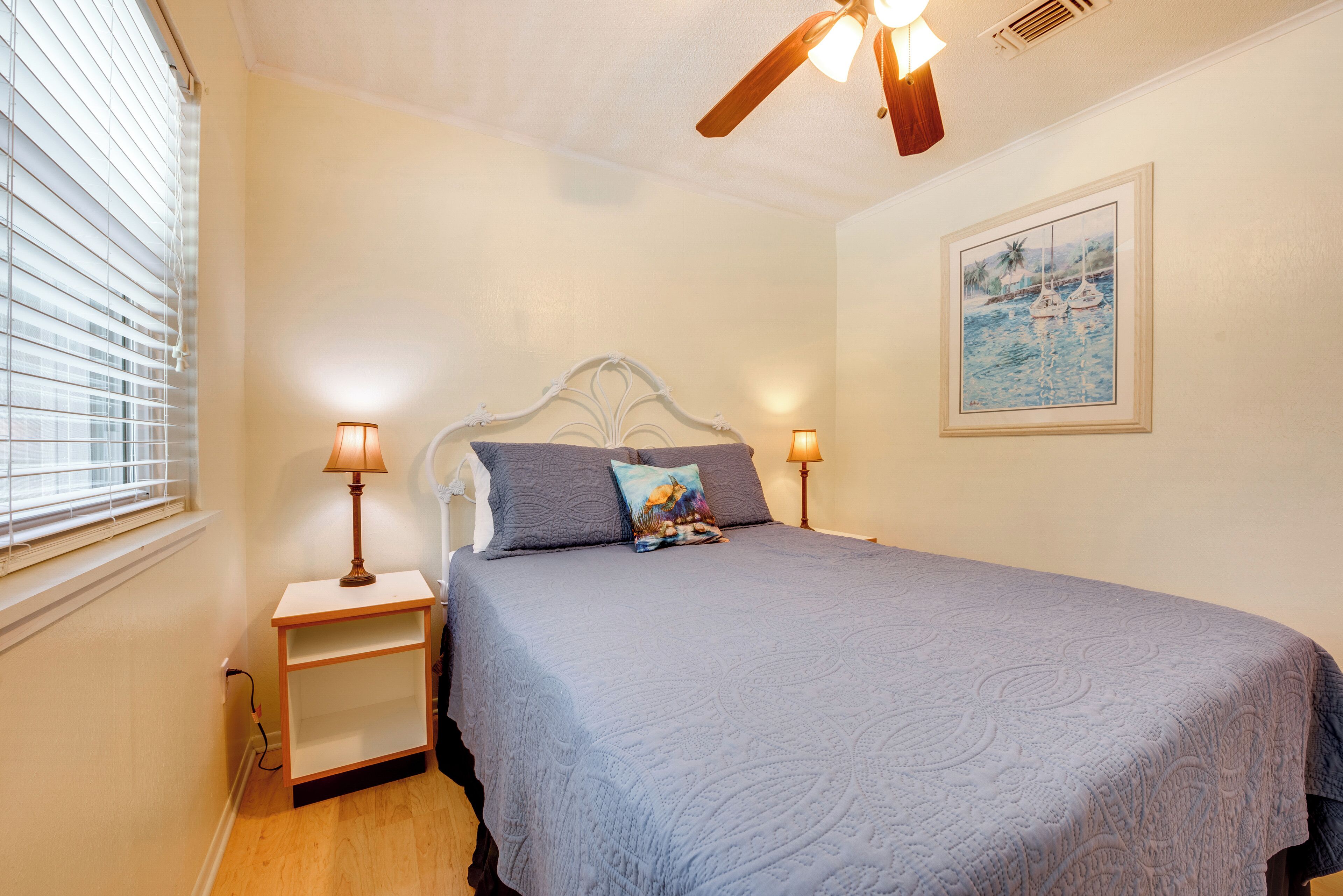 A bedroom with a bed, two bedside lamps, a ceiling fan, a nightstand, and a painting on the wall.