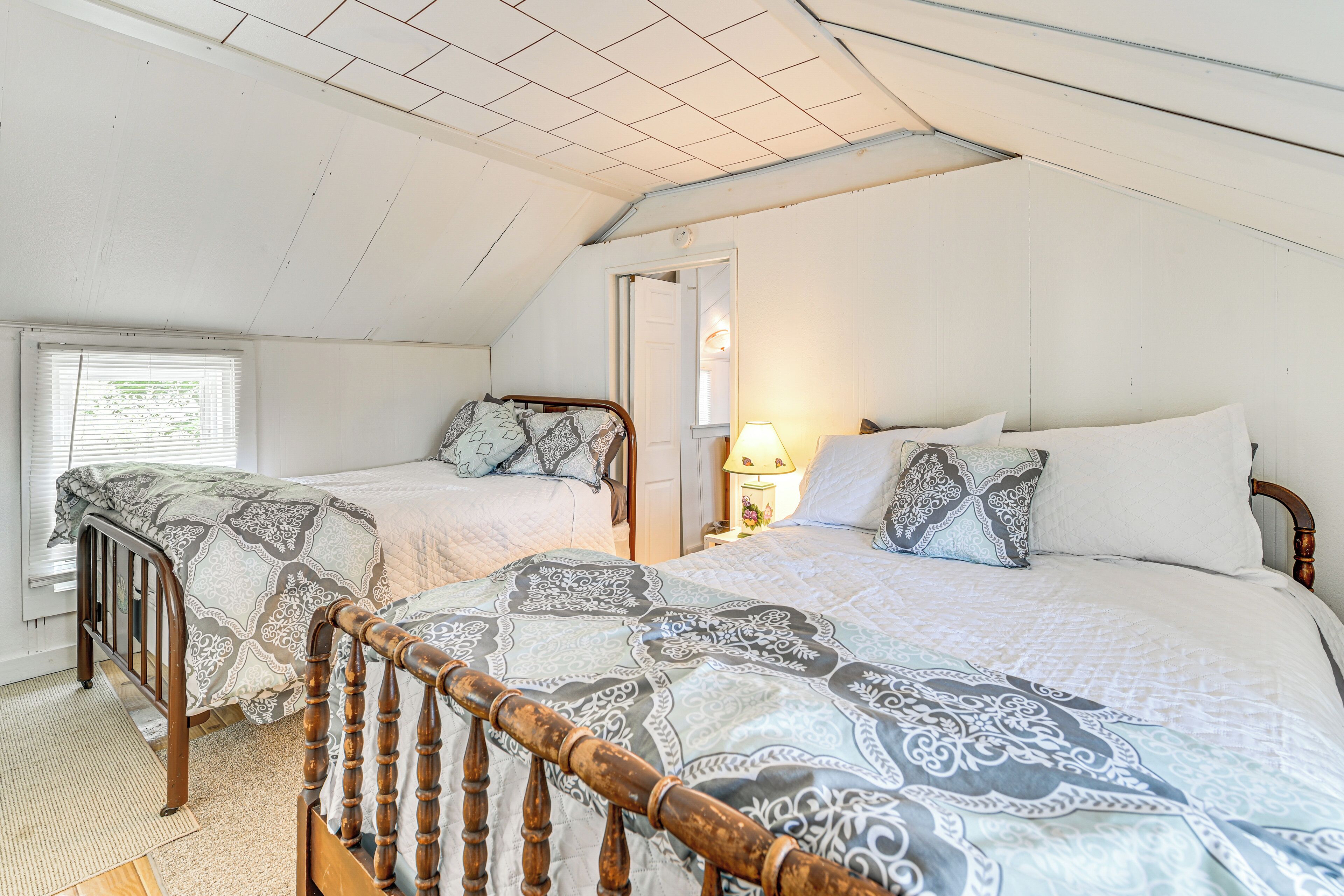 A bedroom with two beds, a wooden headboard, a window with blinds, and a door leading to another room.
