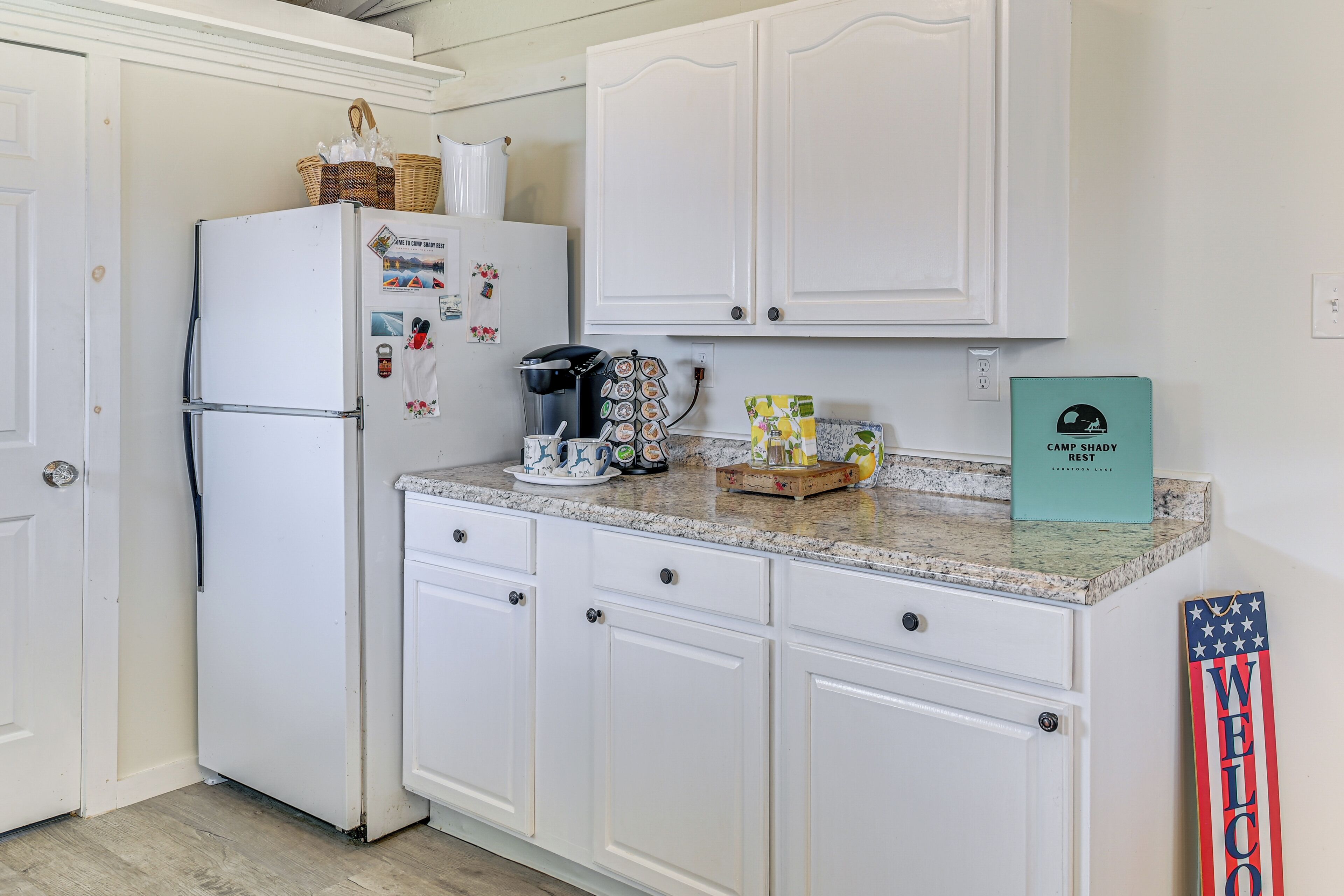 A kitchen with white cabinets, a refrigerator, a coffee maker, and a welcome sign.