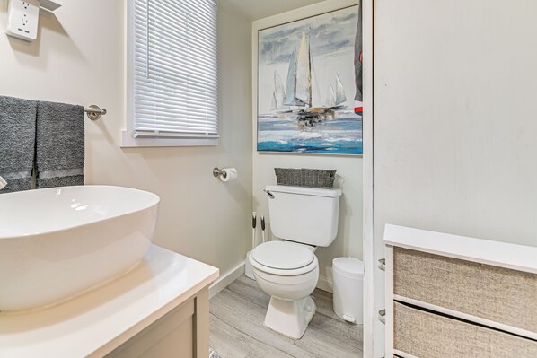 A bathroom with a large white sink, a toilet, a painting of sailboats, and a wicker basket on the toilet tank. - 7 Mi Saratoga Race Course: Lakefront Gem w/ Dock (Saratoga Springs)