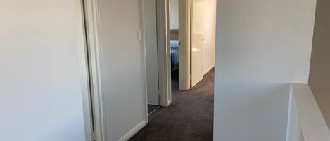 Un corridoio stretto con pareti bianche e pavimento rivestito a moquette, che conduce a una camera da letto con un letto e un comodino.