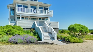 Una casa frente a la playa de varios pisos con una escalera que lleva a un balcón, rodeada de exuberante vegetación y un cielo despejado.