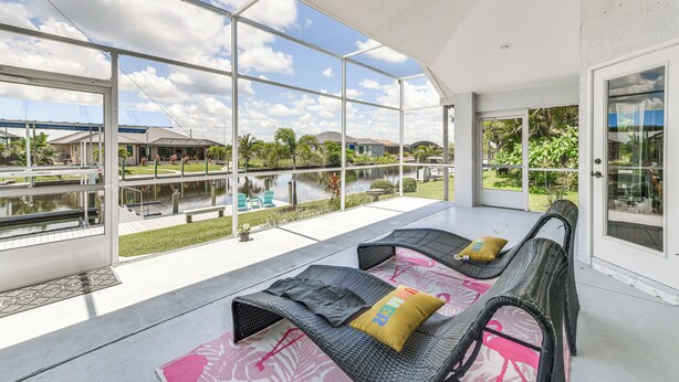 A covered outdoor seating area with wicker furniture and cushions, large glass windows offering views of a lake and surrounding greenery. - 12 Mi to Gulf: Port Charlotte Home w/ Boat Dock (Port Charlotte)