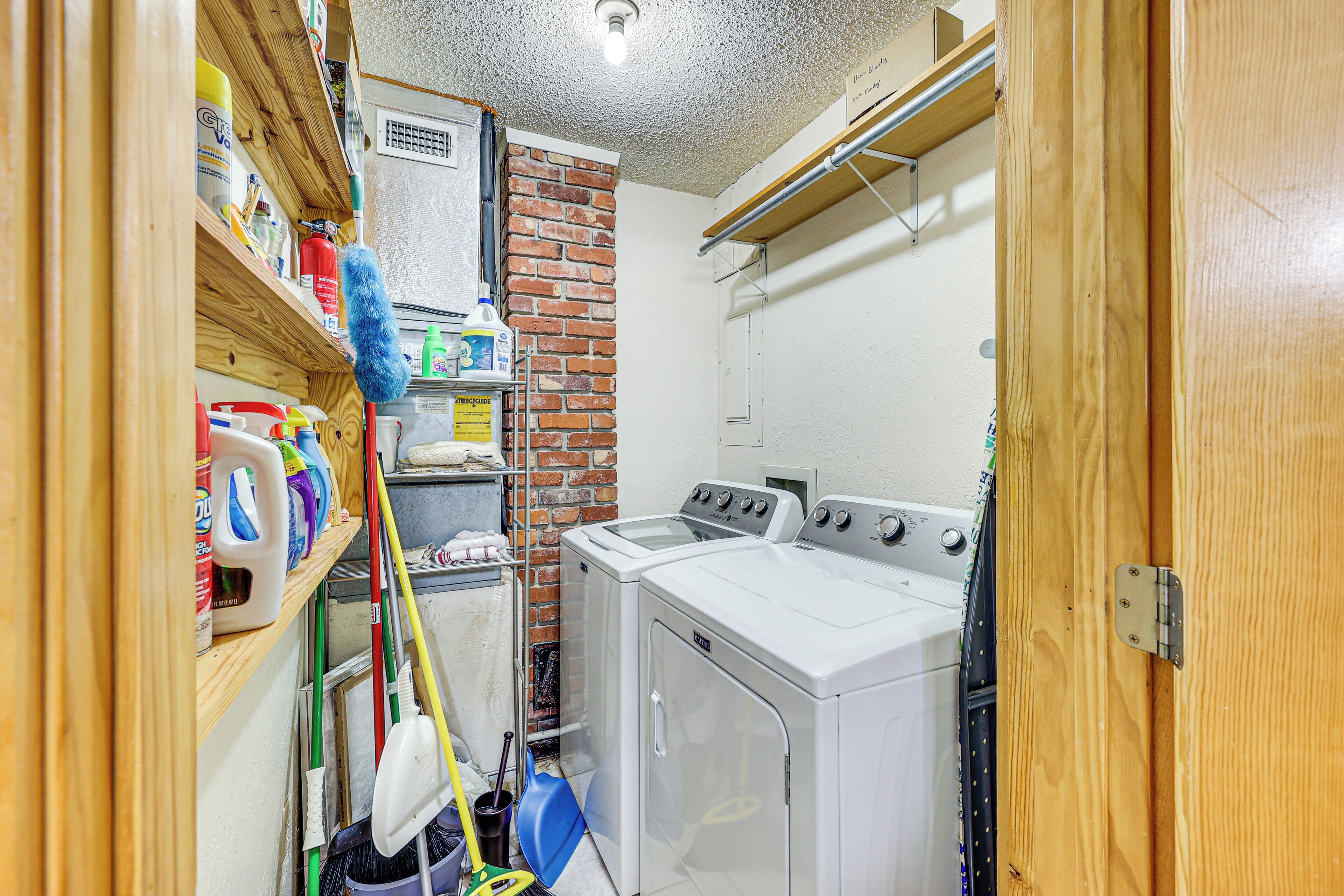 A laundry room with a brick wall, a washing machine, and a dryer. There are cleaning supplies and a mop on the shelves.