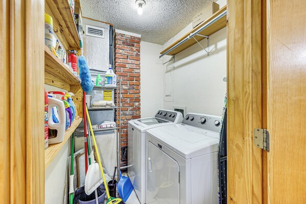 A laundry room with a brick wall, a washing machine, and a dryer. There are cleaning supplies and a mop on the shelves. - < 3 Mi to Broken Bow Lake: Wooded Haven! (Broken Bow)